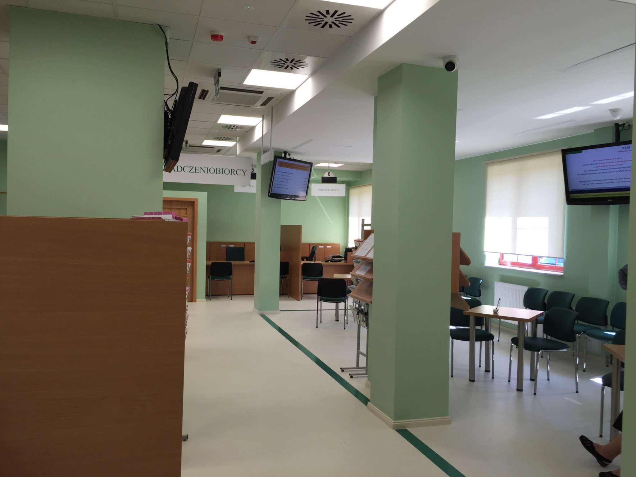 Wnętrze biura z zielonymi ścianami, białymi podłogami z zielonymi akcentami, widoczne biurka, krzesła i ekrany informacyjne.