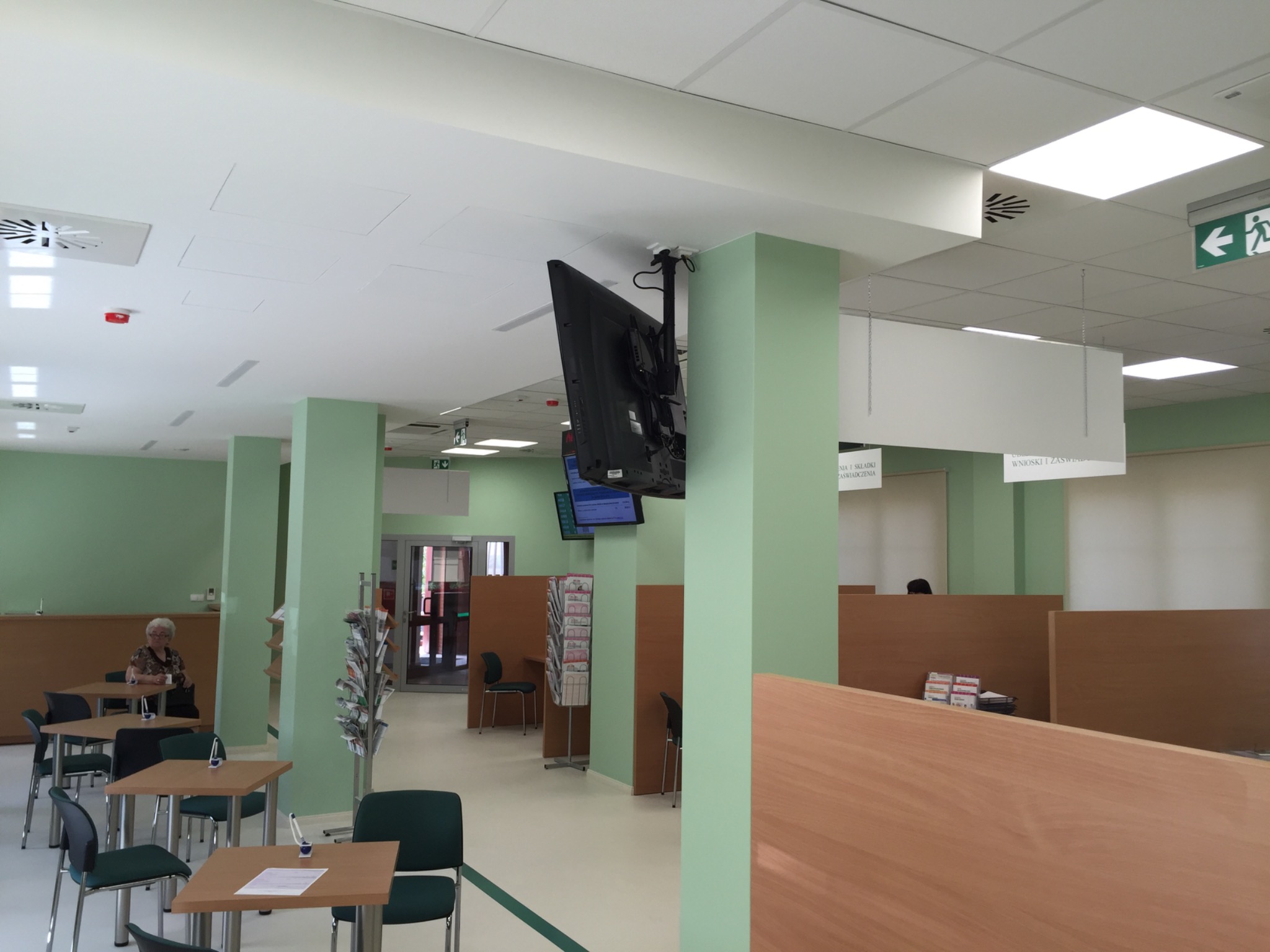 Jasne wnętrze biura z zielonymi kolumnami, drewnianymi przegrodami, stolikami i krzesłami, z wiszącym telewizorem i podwieszanym sufitem.