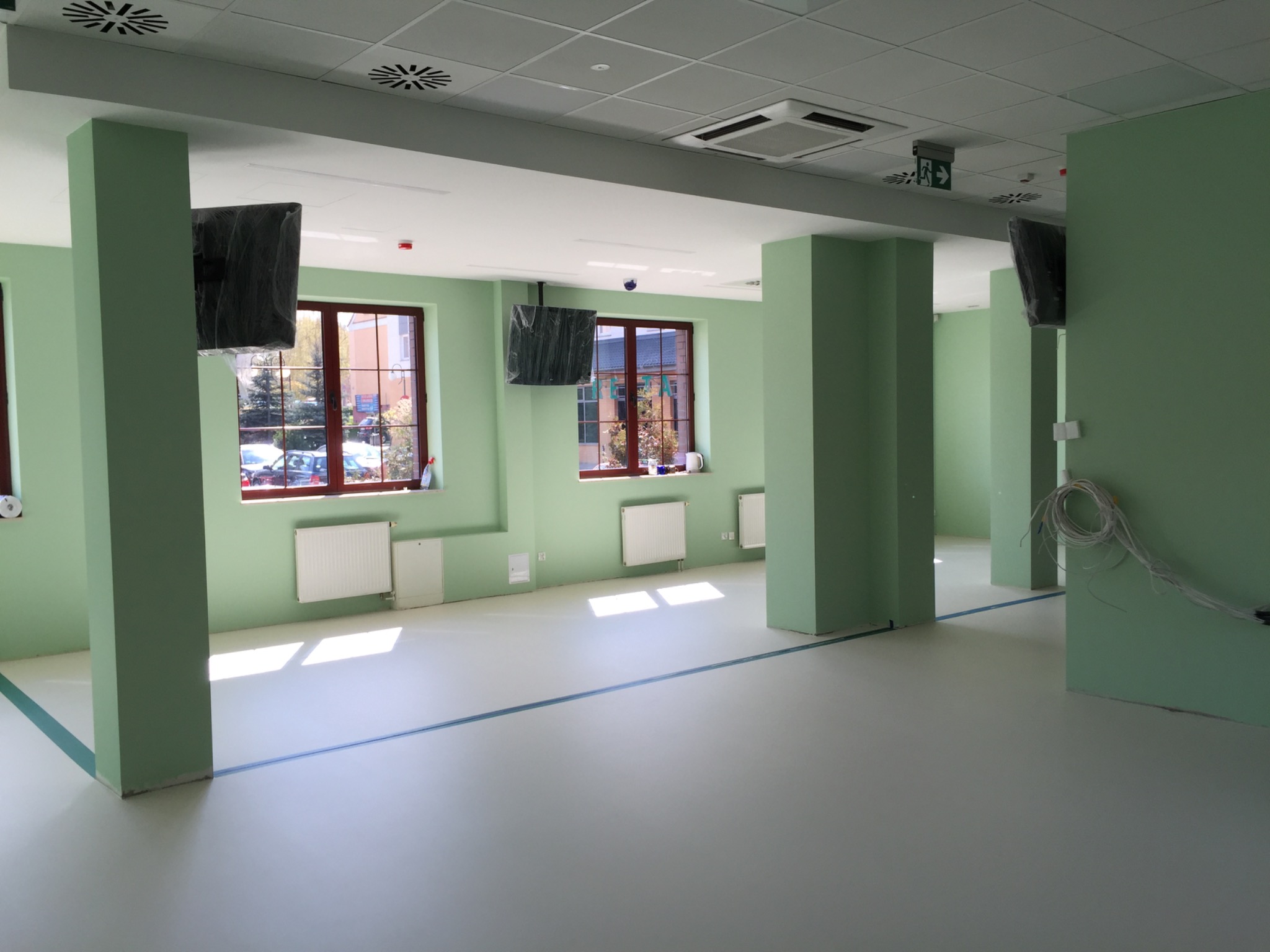 Świeżo wyremontowane biuro w odcieniach zieleni z kolumnami, nową podłogą z zaznaczoną linią, grzejnikami pod oknami i zamontowanymi, ale jeszcze ofoliowanymi telewizorami.