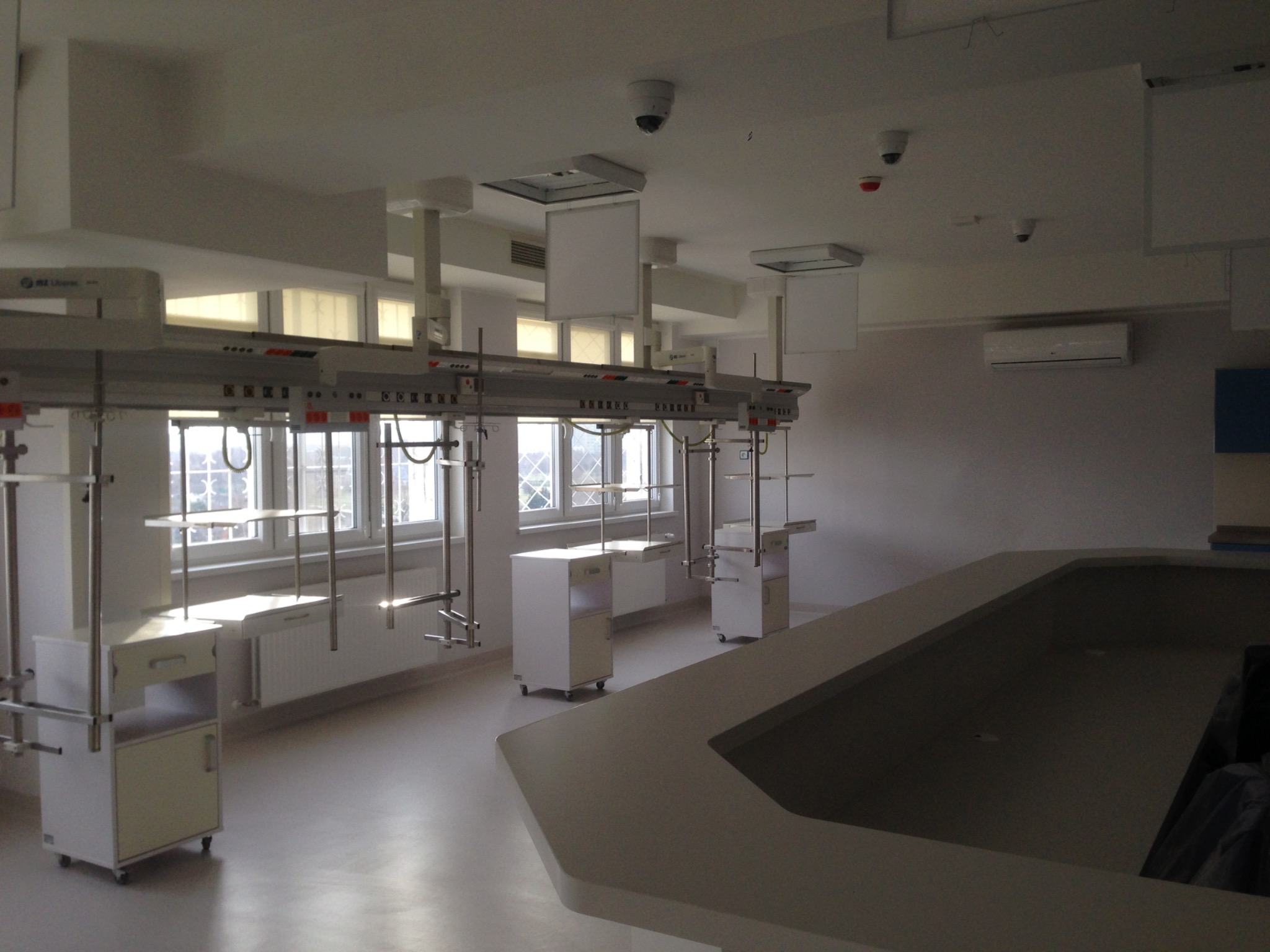 Wnętrze biura lub laboratorium z modułowymi stanowiskami pracy: białe szafki na kółkach, metalowe stelaże podwieszane do sufitu, okna z roletami, klimatyzacja i kamery przemysłowe