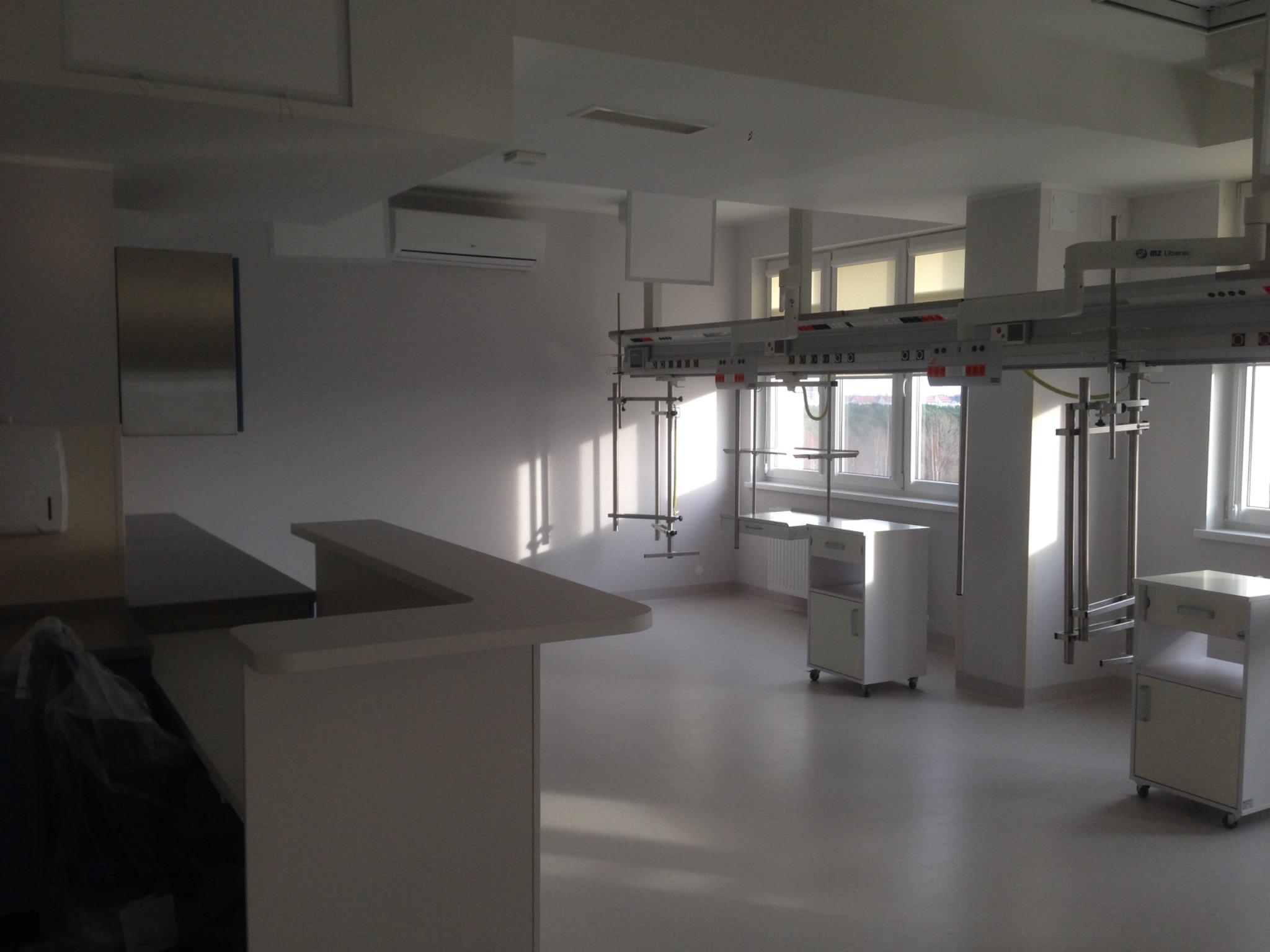 Jasne, przestronne wnętrze biurowe z elementami wyposażenia medycznego, wpadające promienie słoneczne podkreślają czystość pomieszczenia, widoczne szafki na kółkach i metalowe konstrukcje pod sufitem.