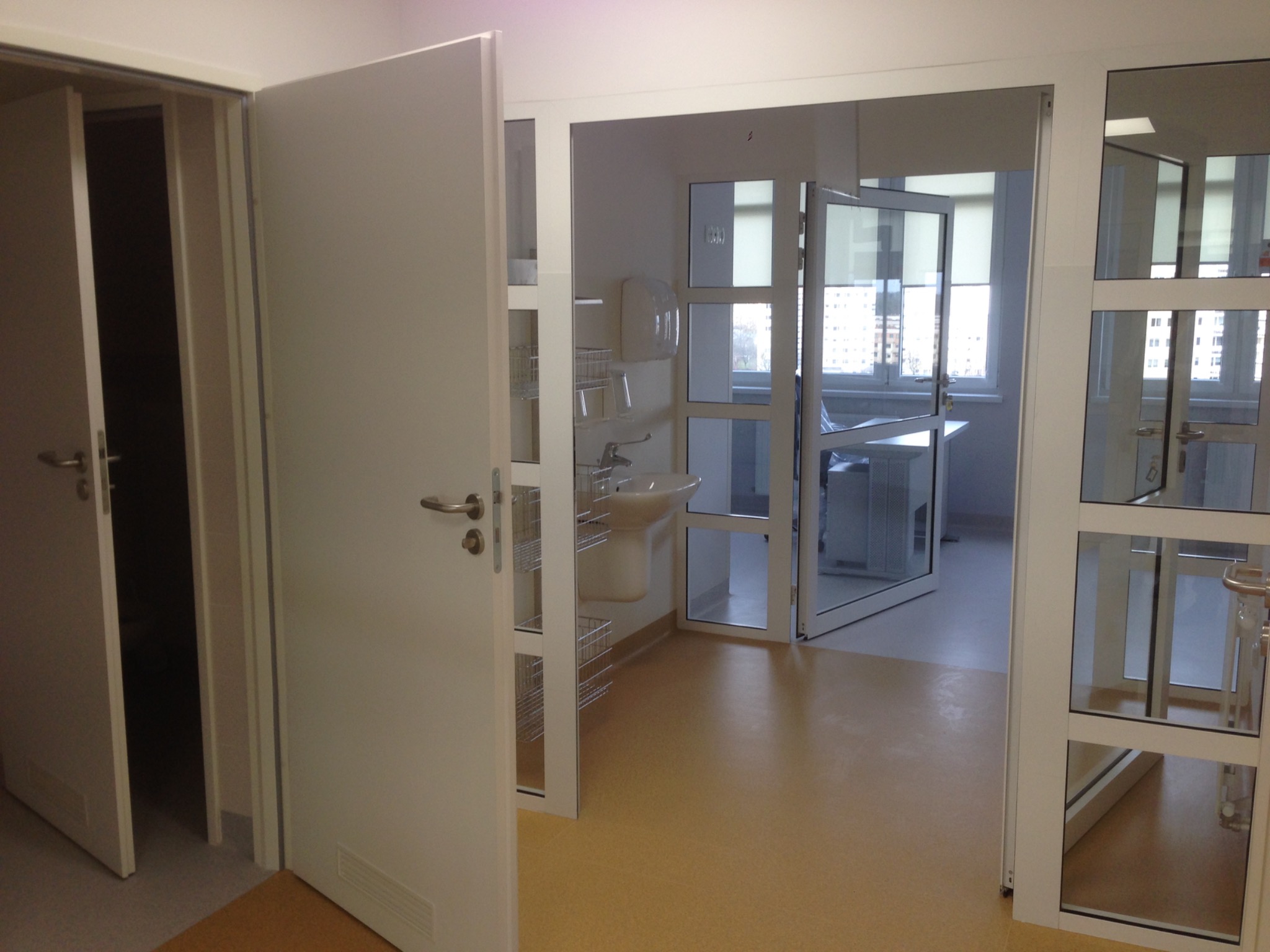 Wnętrze biura lub placówki medycznej z widoczną umywalką, metalowymi koszami, jasną podłogą i przeszklonymi ścianami działowymi, widziane przez otwarte drzwi.