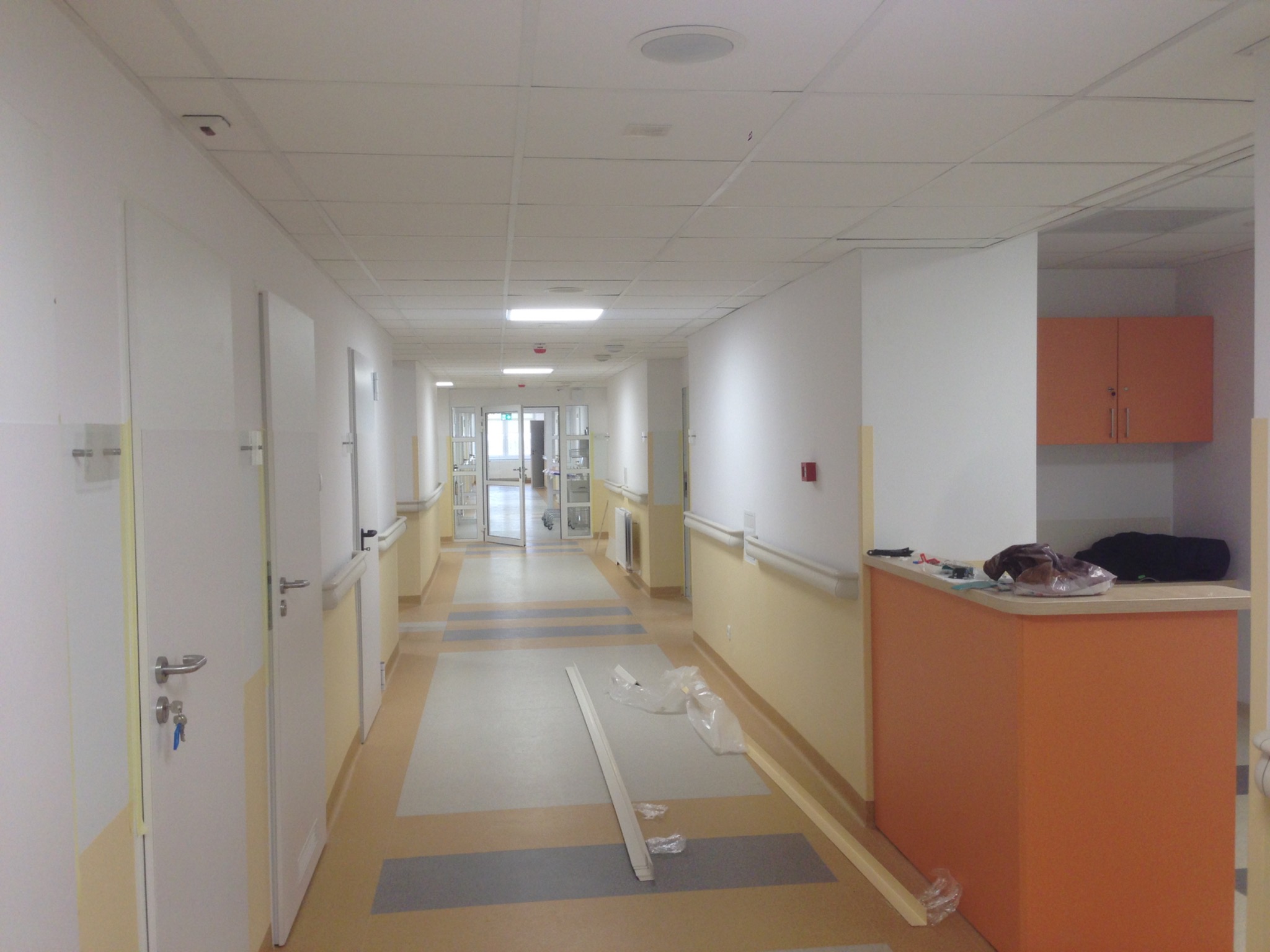 Długi korytarz w budynku biurowym w trakcie malowania, widoczne świeżo położone panele podłogowe, pomalowane ściany na biało i żółto, białe drzwi z srebrnymi klamkami i kluczami w zamku...