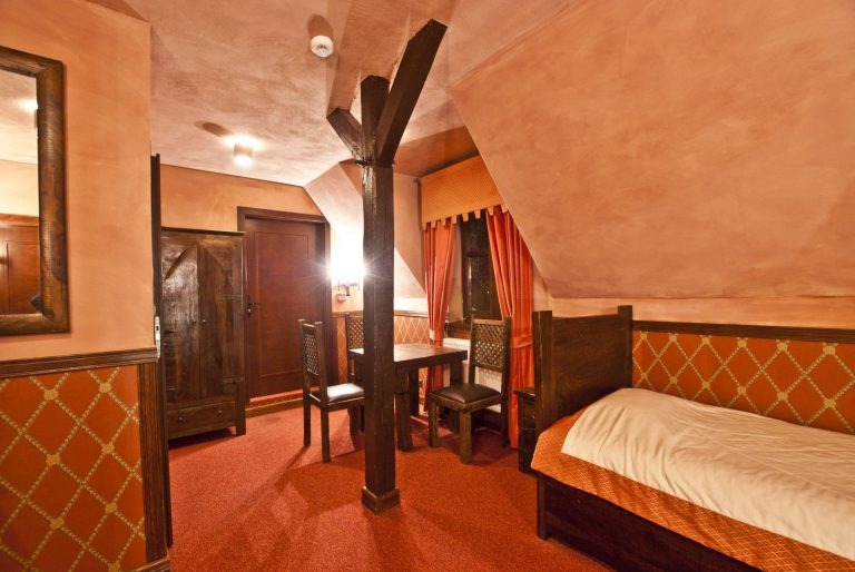 Rustykalny pokój hotelowy na poddaszu z drewnianą belką podtrzymującą strop, meblami w ciemnym drewnie, czerwonym dywanem i pomarańczowymi ścianami.