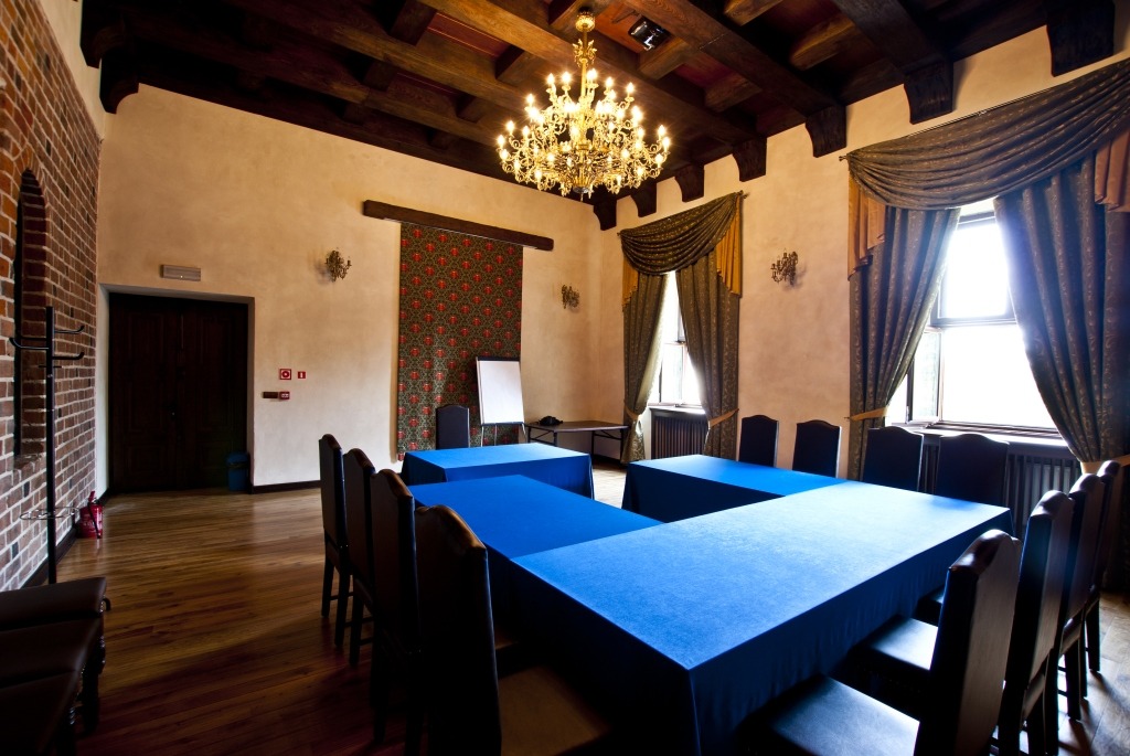 Sala konferencyjna w stylu pałacowym z drewnianym stropem, kryształowym żyrandolem, oknami z ozdobnymi zasłonami i stołami nakrytymi niebieskim obrusem w układzie podkowy.
