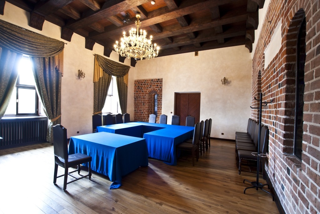 Sala konferencyjna w stylu zamkowym z drewnianym stropem, kryształowym żyrandolem, oknami z ciężkimi zasłonami i stołami przykrytymi niebieskimi obrusami.