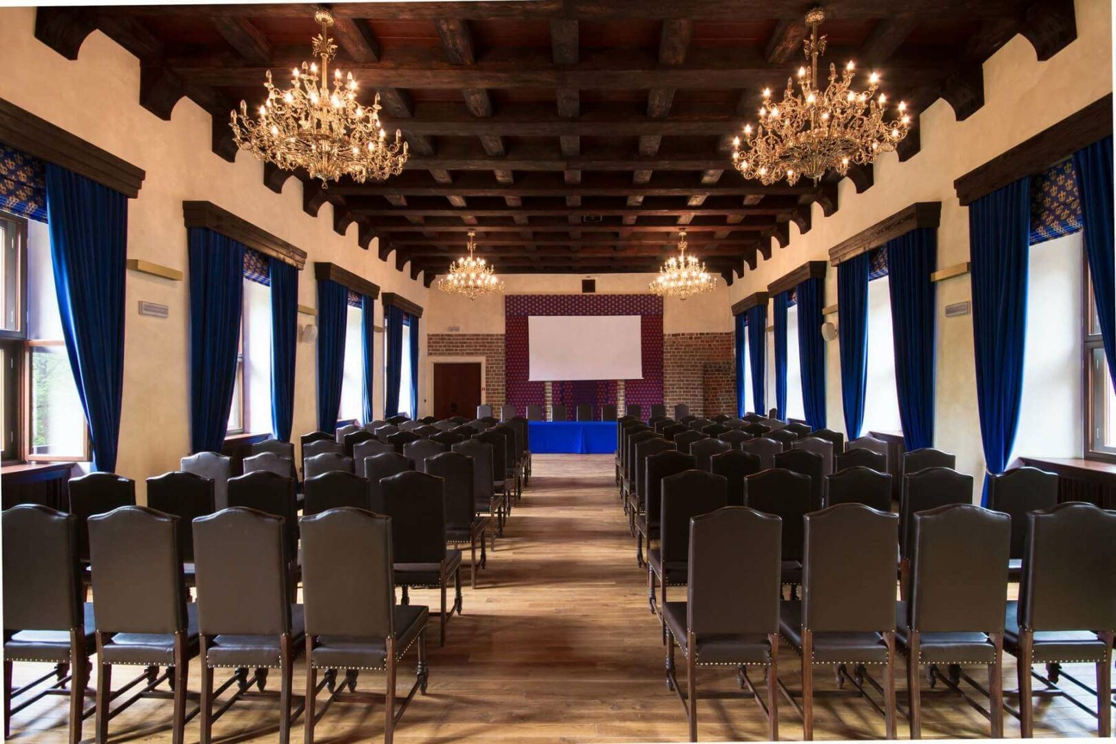 Sala Rycerska przygotowana na konferencję: rzędy krzeseł ustawione w kierunku ekranu projekcyjnego, z drewnianym stropem belkowym i ozdobnymi żyrandolami