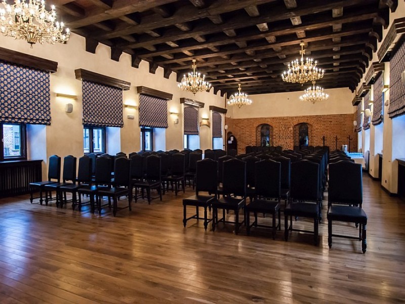 Sala rycerska z drewnianym stropem i parkietem, rzędy czarnych krzeseł ustawione do ceremonii, kryształowe żyrandole i okna z roletami w kolorze niebieskim.
