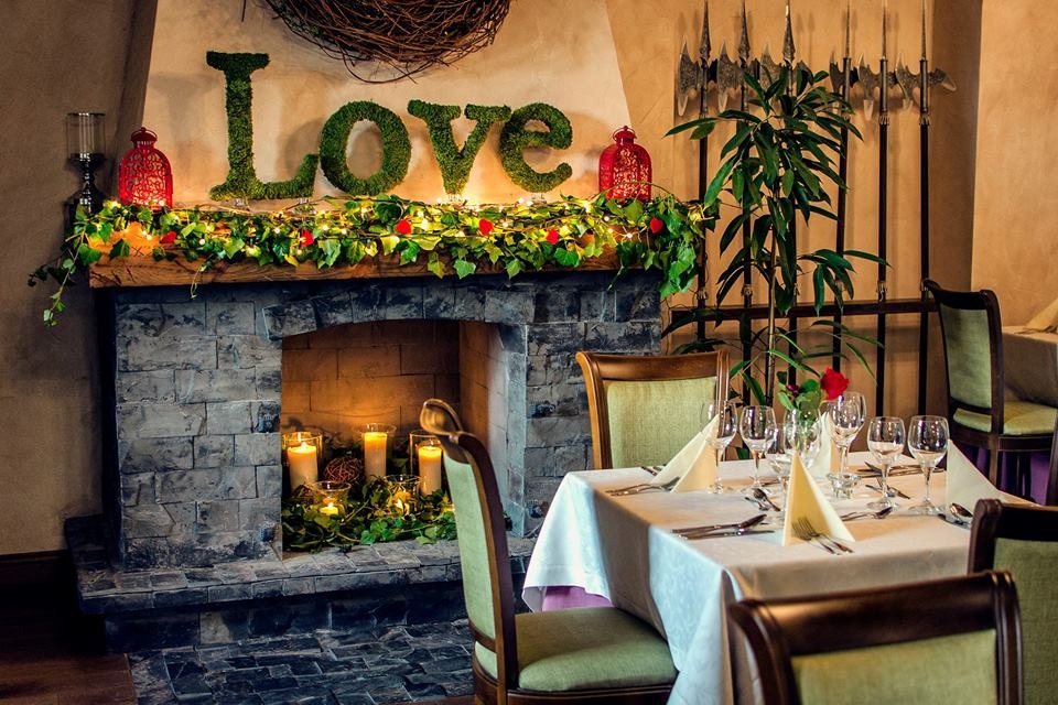 Uroczysta dekoracja sali z kamiennym kominkiem, nad którym widnieje napis LOVE z mchu, oświetlony girlandą, oraz nakryty stół z kieliszkami i serwetkami.