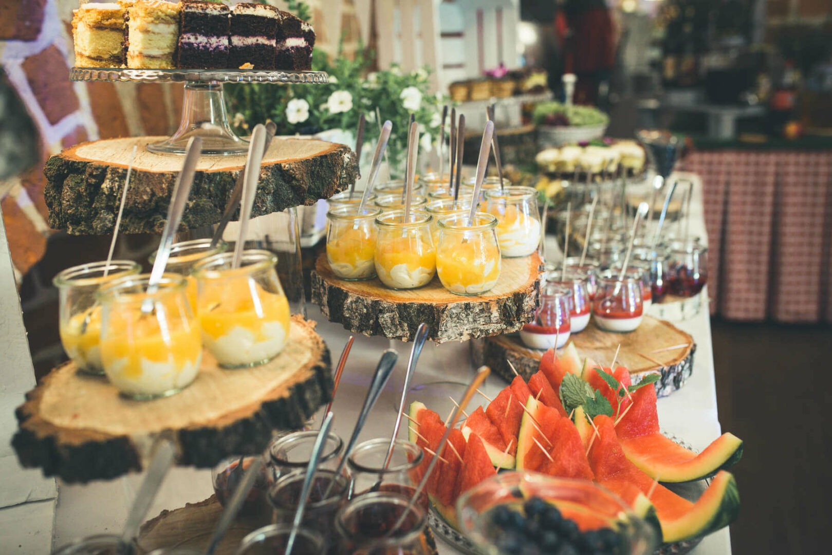 Stół z deserami na drewnianych podstawkach: ciasta, musy w słoiczkach, arbuz na patykach, jagody.