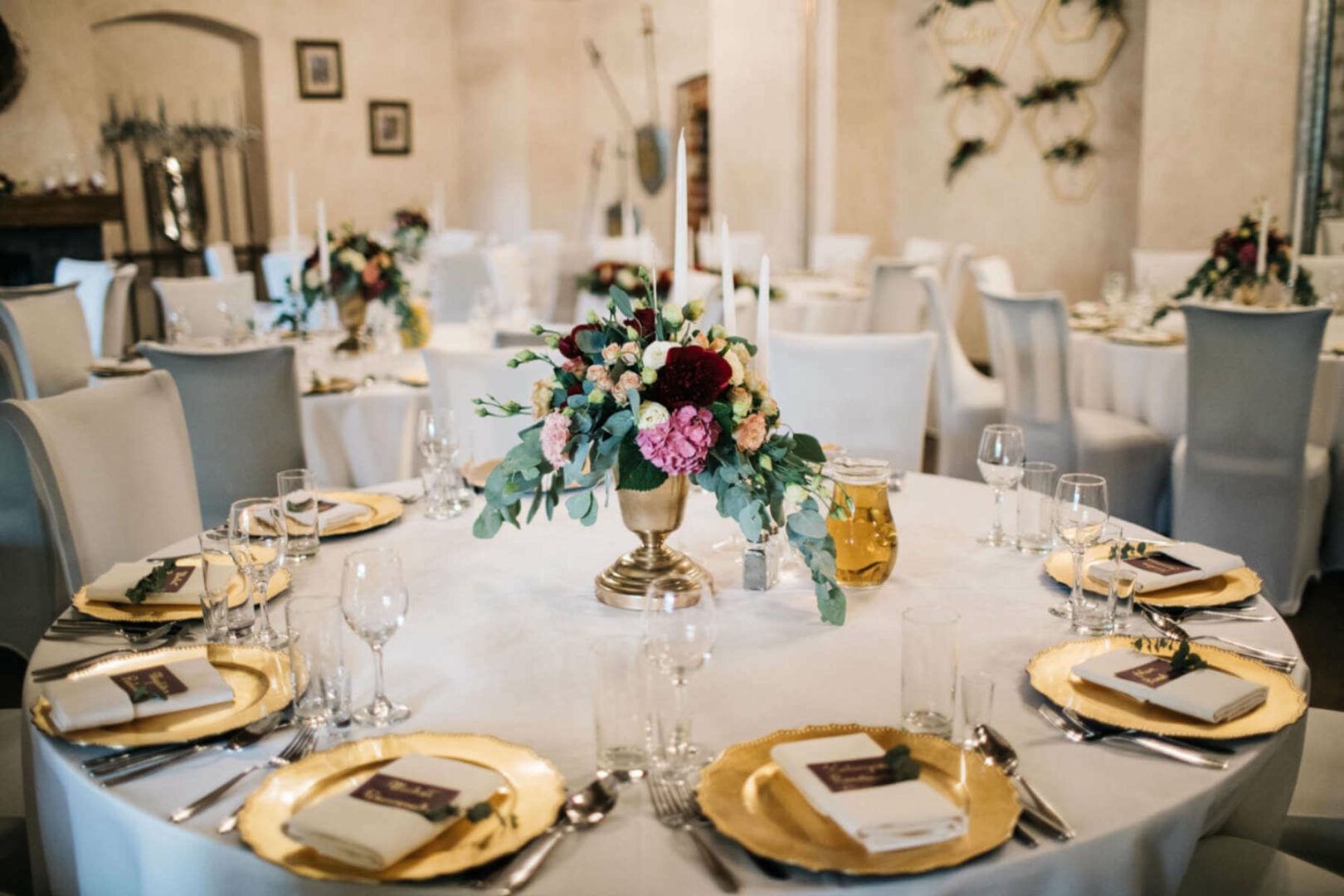 Elegancko nakryty stół weselny z kwiatową dekoracją w złotej wazie, złote talerze i białe serwetki z bordowymi etykietami.