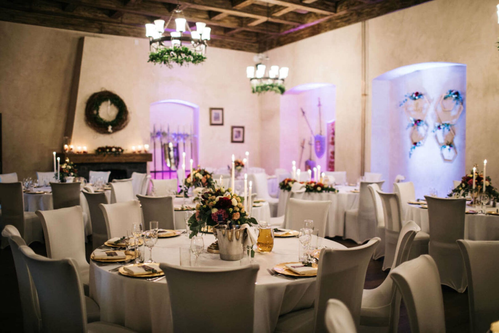 Uroczysta sala bankietowa z okrągłymi stołami, białymi krzesłami, kwiatowymi dekoracjami, świecami i nastrojowym oświetleniem w odcieniach fioletu.