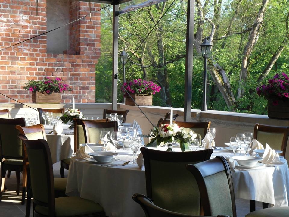 Uroczysty stół z białym obrusem, nakryty zastawą i ozdobiony kwiatami, ustawiony na zewnątrz z widokiem na zieleń, dekorowany skrzynkami z fioletowymi kwiatami.