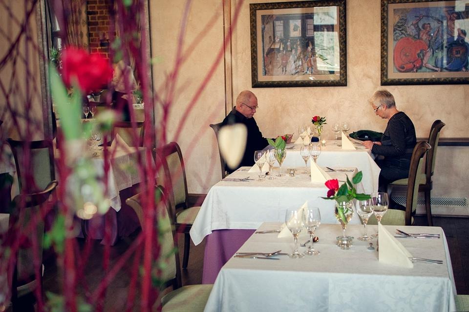 Elegancko nakryte stoły w restauracji, z dwoma osobami czytającymi menu przy stolikach, dekoracje z czerwonych kwiatów w wazonach.