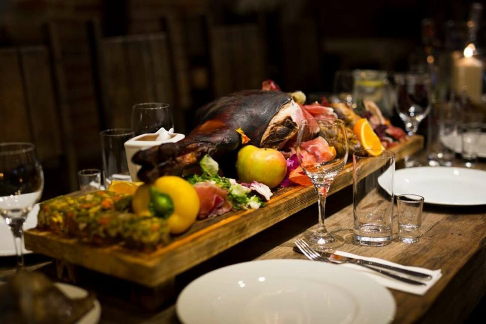 Uroczysty stół z pieczoną nogą, wędlinami, owocami i warzywami na drewnianej desce, przygotowany do celebracji.