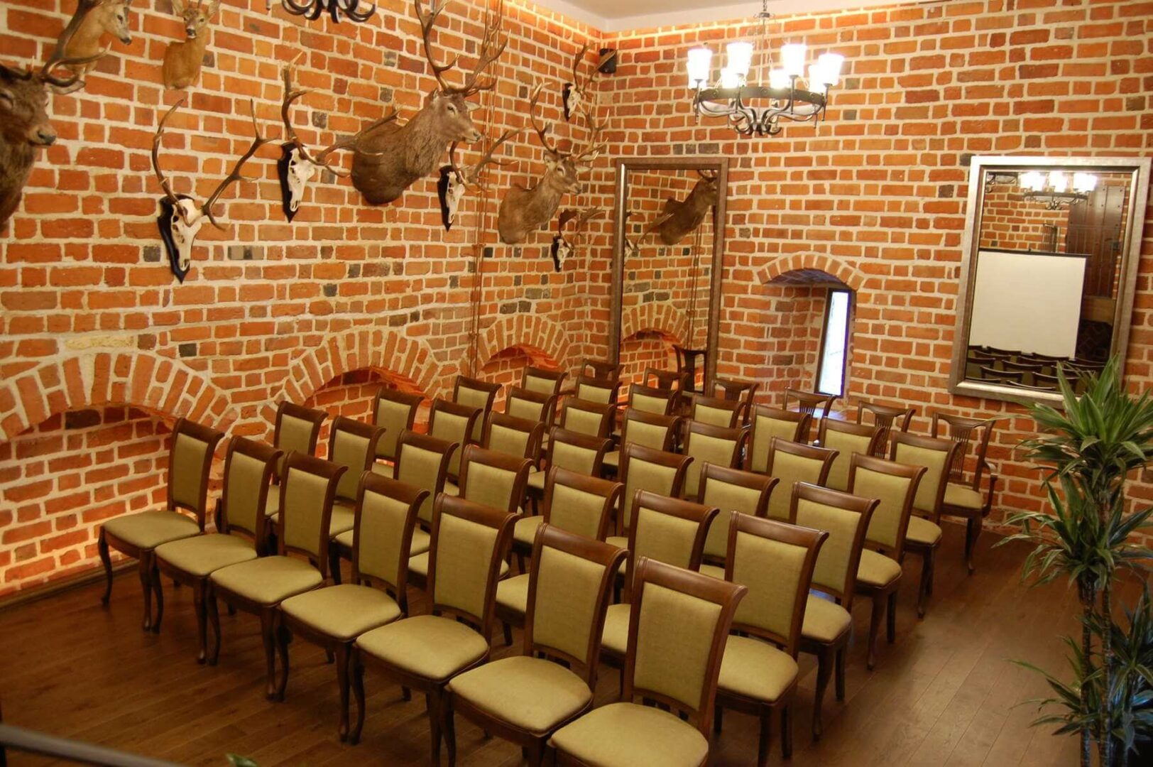 Sala konferencyjna z rzędami krzeseł na drewnianej podłodze, ceglane ściany zdobione trofeami myśliwskimi i lustrami, oświetlona żyrandolem.