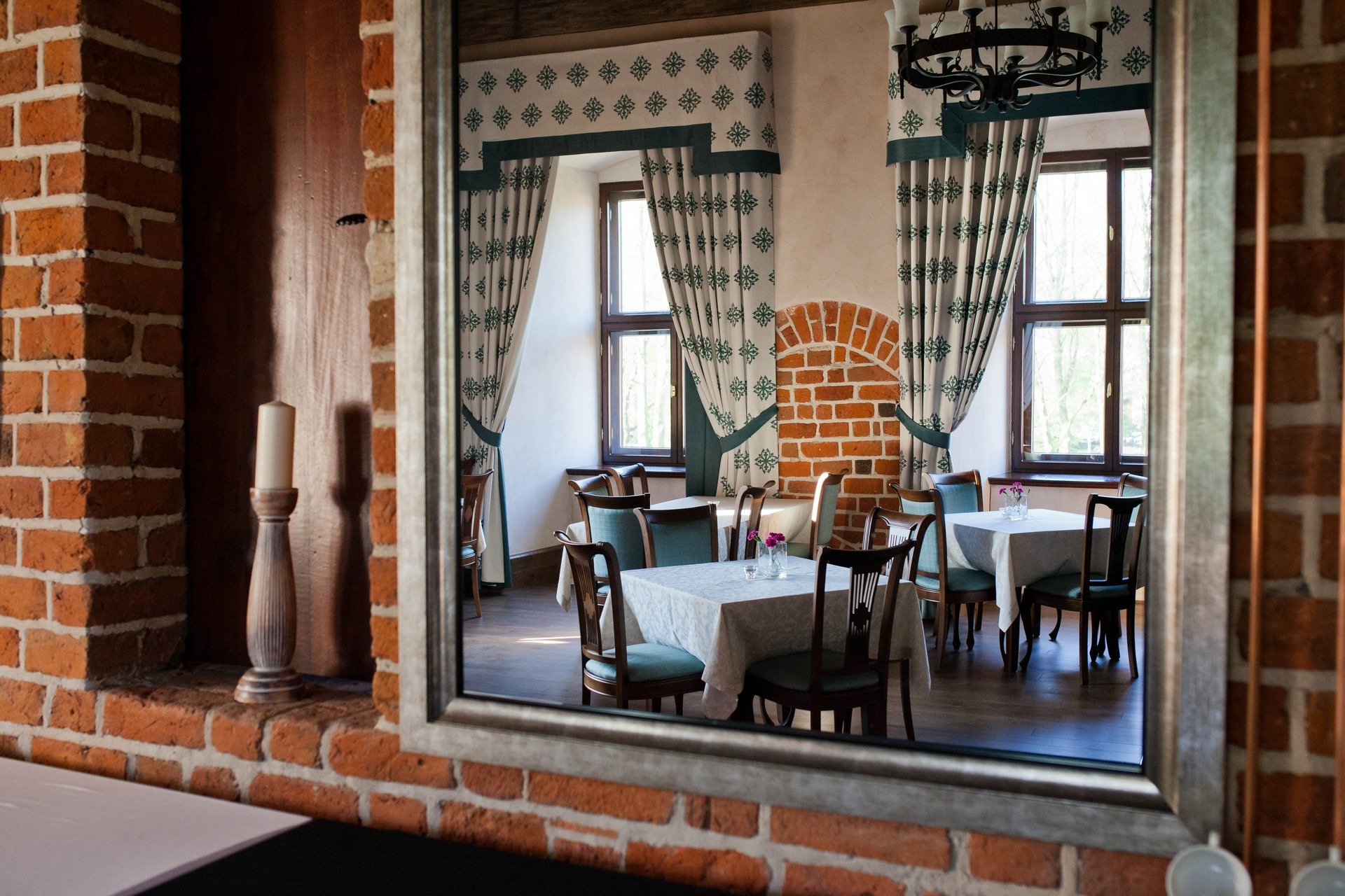 Uroczyste wnętrze restauracji widziane w lustrze, z ceglanymi elementami, eleganckimi zasłonami w kwiaty i stolikami nakrytymi białymi obrusami, z widoczną świecą na ceglanej półce po lewej stronie.