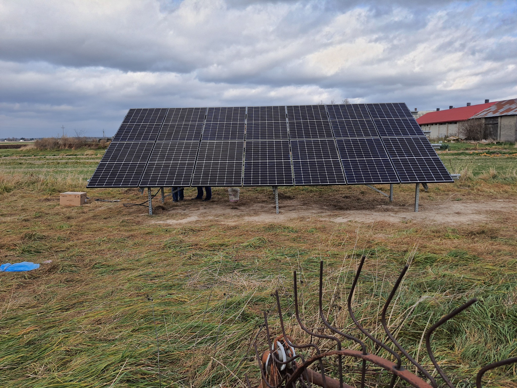 Montaż paneli fotowoltaicznych na polu w pochmurny dzień. W tle widoczne budynki gospodarcze z czerwonym dachem. Na pierwszym planie zardzewiałe widły.