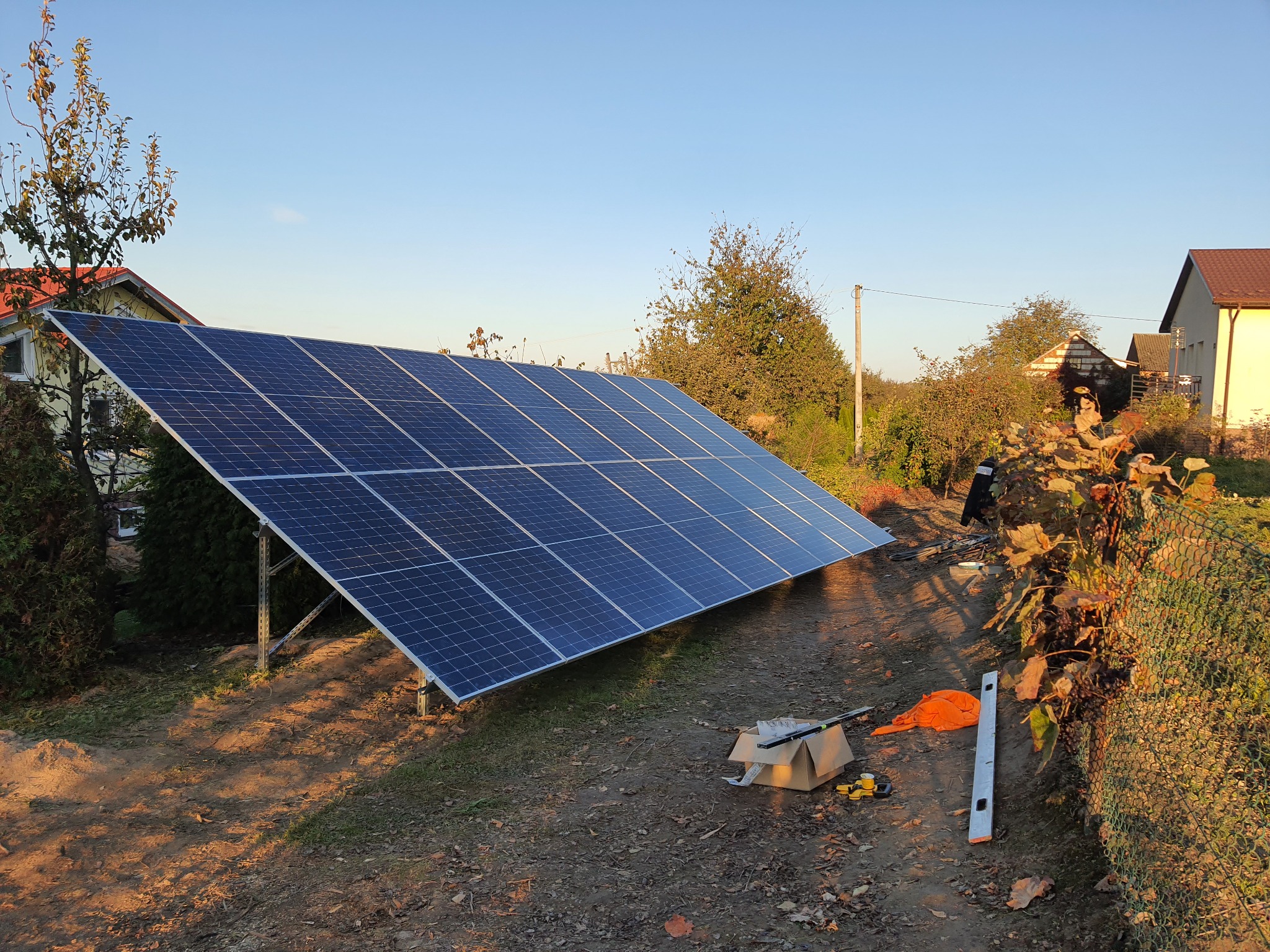 Instalacja paneli słonecznych na gruncie, widoczne narzędzia pracy, poziomica, karton i pomarańczowa torba, w tle domy jednorodzinne i drzewa.