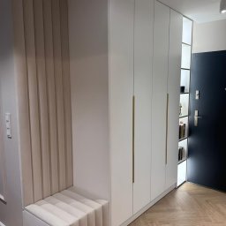 WOODMEBLO Michał Steczko - Biała szafa wnękowa z siedziskiem obitym beżową tkaniną w przedpokoju. Złote uchwyty, parkiet w jodełkę i czarne drzwi wejściowe.