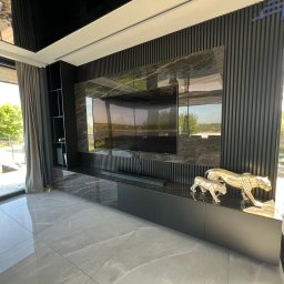 WOODMEBLO Michał Steczko - Nowoczesna zabudowa TV z czarnymi lamelami i marmurowym akcentem, uzupełniona o dekoracyjne figurki panter. Minimalistyczny design w luksusowym wnętrzu.