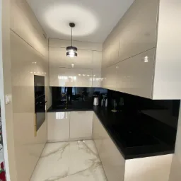 Kompaktowa kuchnia w odcieniach beżu z czarnym blatem i złotymi akcentami, oświetlona nowoczesną lampą wiszącą. Widoczna płyta indukcyjna i armatura.