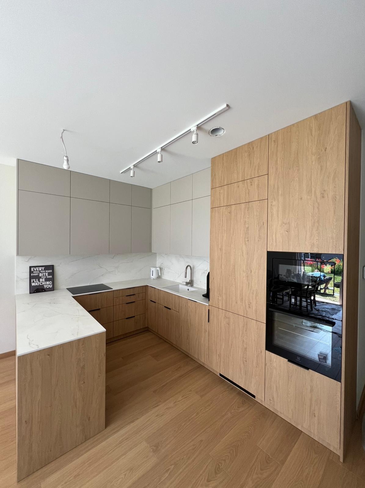 Jasna kuchnia w kształcie litery U z drewnianymi frontami i białym blatem. Na blacie czarna płyta indukcyjna i dekoracyjny napis. Oświetlenie szynowe.