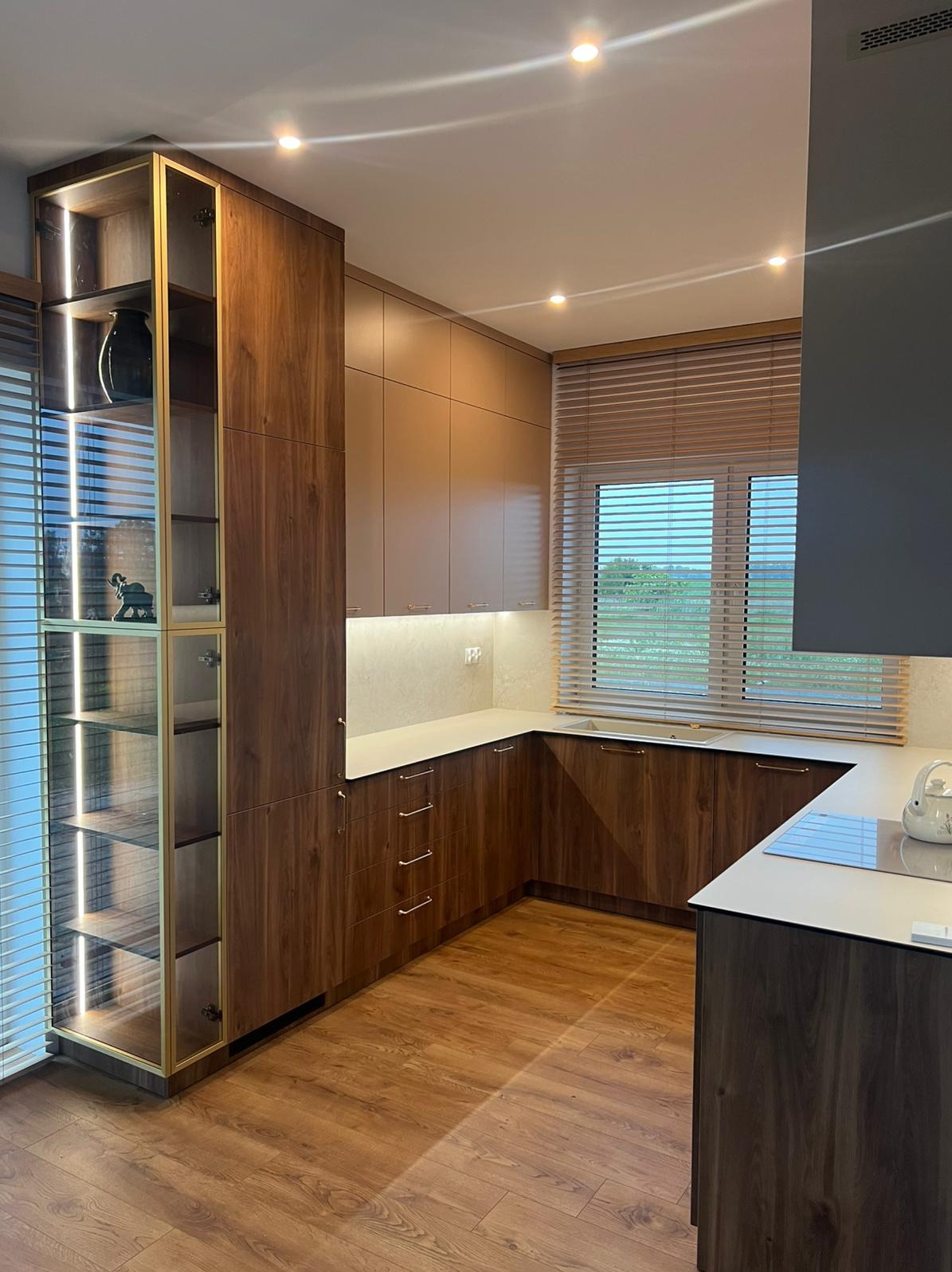 Nowoczesna kuchnia z drewnianymi frontami, oświetleniem LED i przeszkloną witryną. Jasny blat i drewniana podłoga tworzą ciepłą atmosferę.