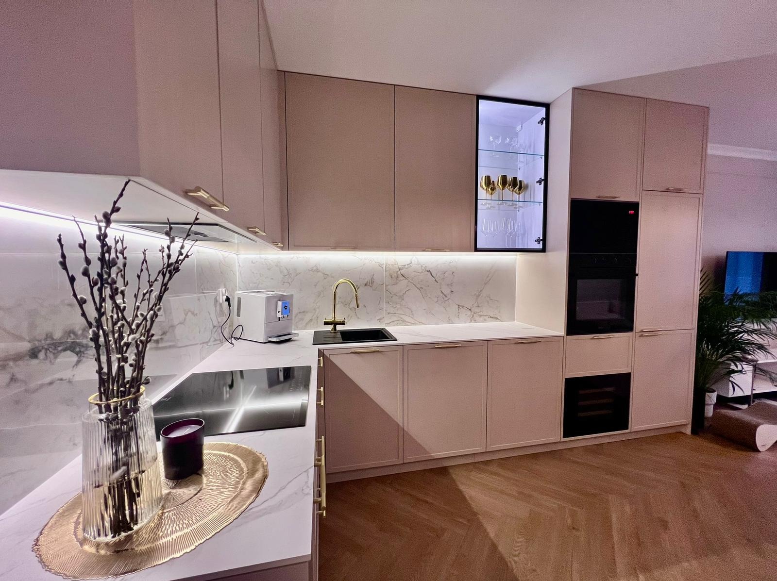 Beżowa kuchnia z marmurowym blatem i złotymi akcentami: armatura, uchwyty, dekoracje. Podświetlane szafki i blat. Na blacie ekspres do kawy i wazon z gałązkami.