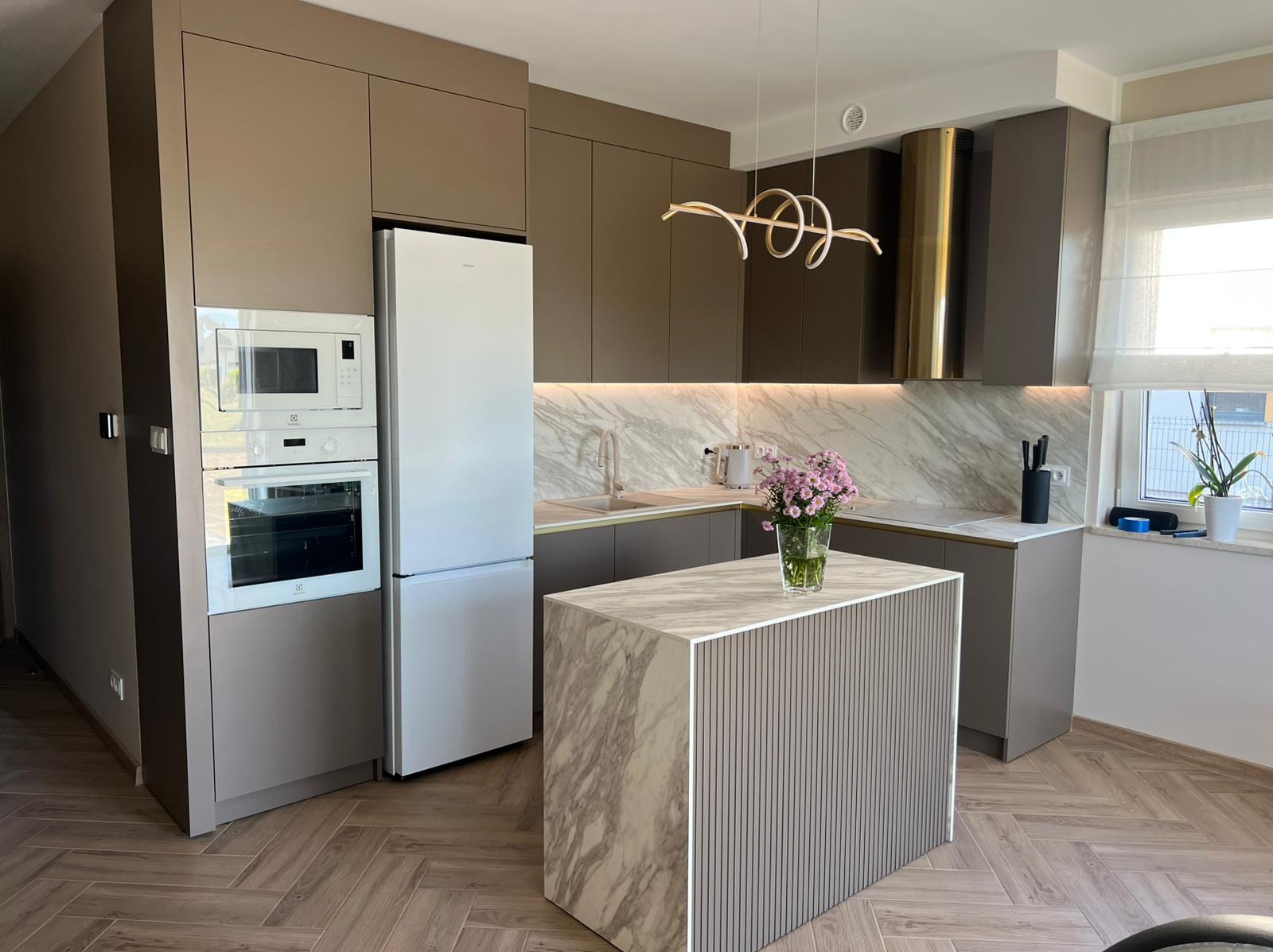 Nowoczesna kuchnia w odcieniach szarości i beżu z wyspą, marmurowym blatem i złotymi akcentami. Oświetlenie LED podkreśla elegancki design. Parkiet w jodełkę.