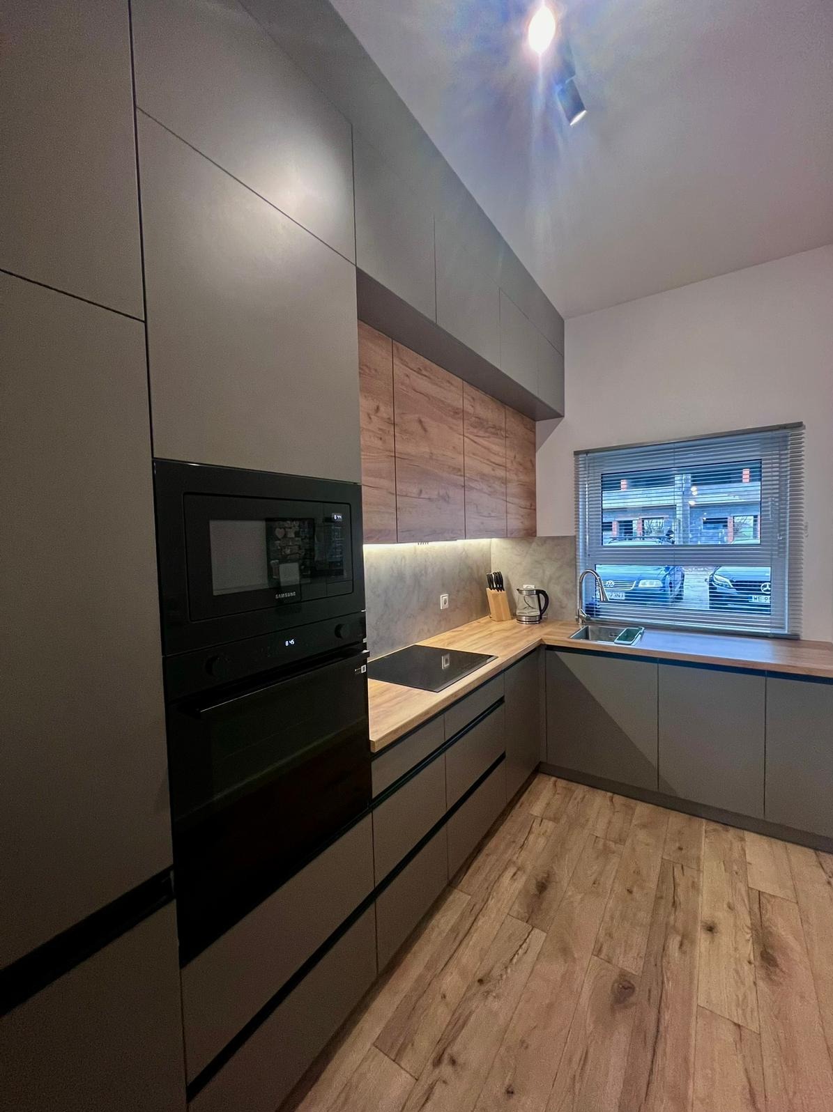 Szara kuchnia z drewnianymi akcentami: zabudowa z piekarnikiem i mikrofalówką, drewniany blat, okno z żaluzjami, drewniana podłoga. Nowoczesny design.