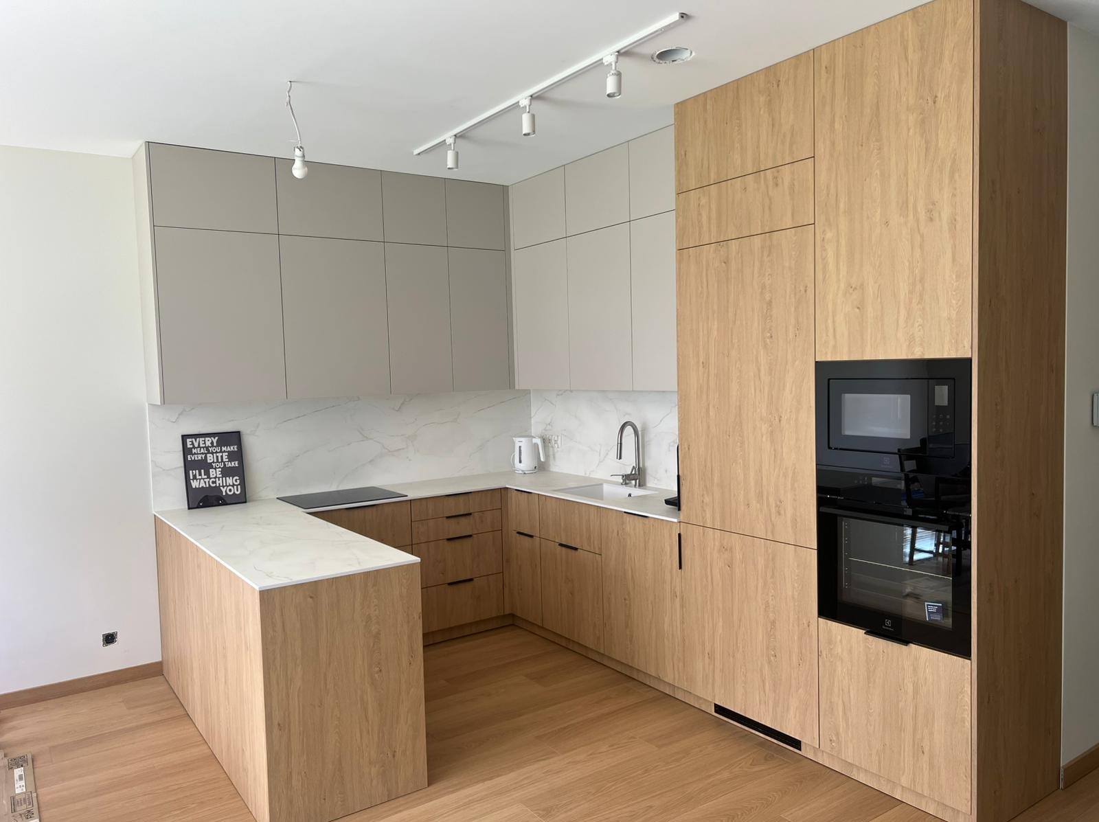 Jasna kuchnia w kształcie litery U z drewnianymi frontami i szarymi szafkami wiszącymi, marmurowy blat i backsplash, oświetlenie punktowe.