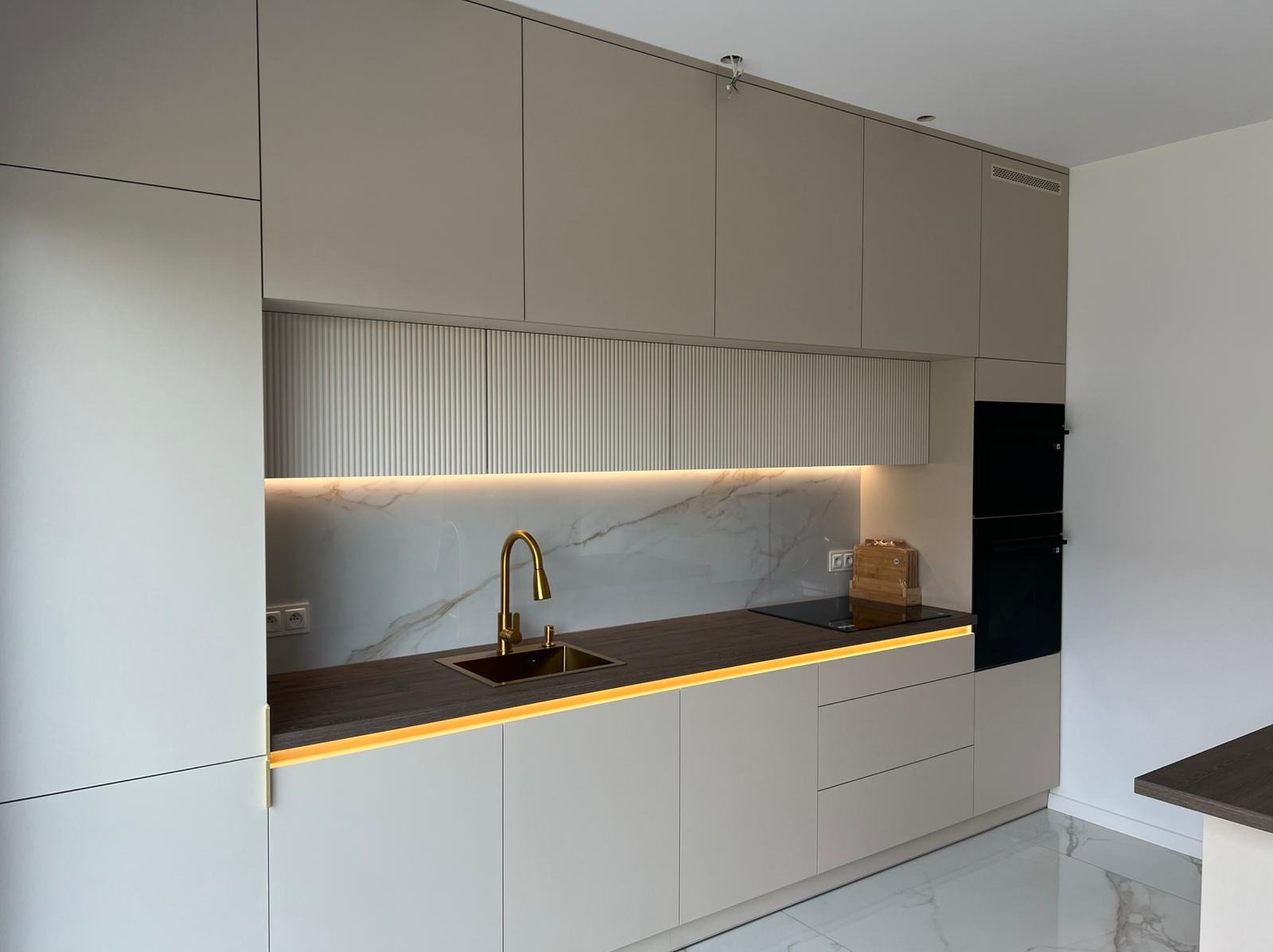 Nowoczesna kuchnia z beżowymi frontami, marmurowym backsplashiem i złotą armaturą. Podświetlenie LED podkreśla blat i dodaje elegancji. Widoczny piekarnik w zabudowie.