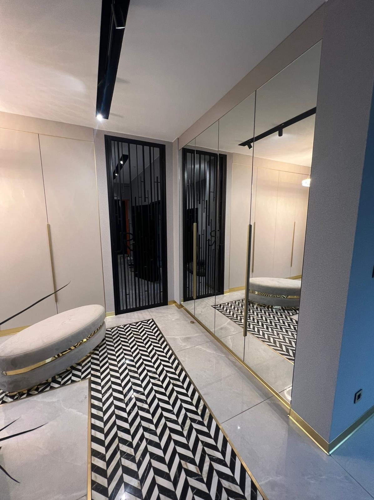 Nowoczesna szafa wnękowa z lustrzanymi drzwiami i złotymi akcentami w Jasin. Geometryczny wzór dywanu i minimalistyczne oświetlenie podkreślają elegancję wnętrza.