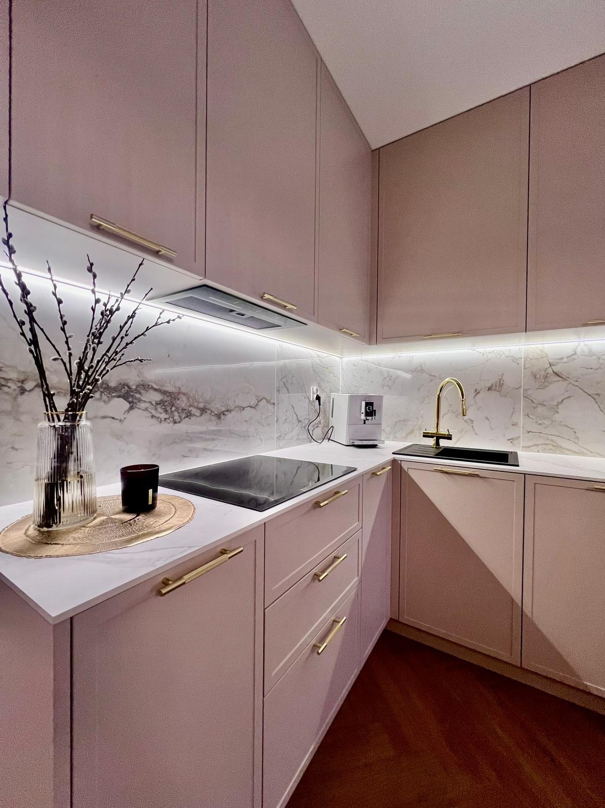 Różowa kuchnia z marmurowym backsplashiem, złotymi uchwytami i oświetleniem LED pod szafkami. Na blacie wazon z gałązkami, płyta indukcyjna i ekspres do kawy.