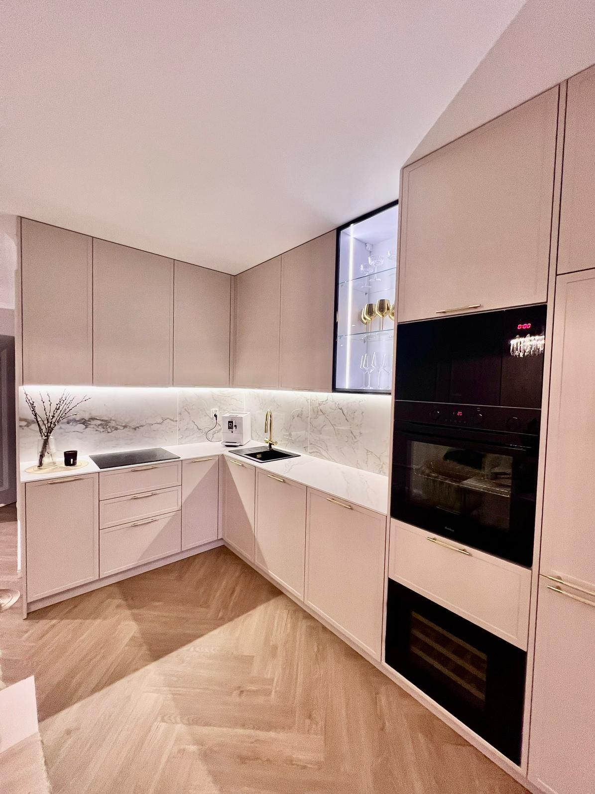 Jasna kuchnia z marmurowym blatem i złotymi akcentami. W tle podświetlana witryna z kieliszkami. Nowoczesny design z drewnianą podłogą w jodełkę.