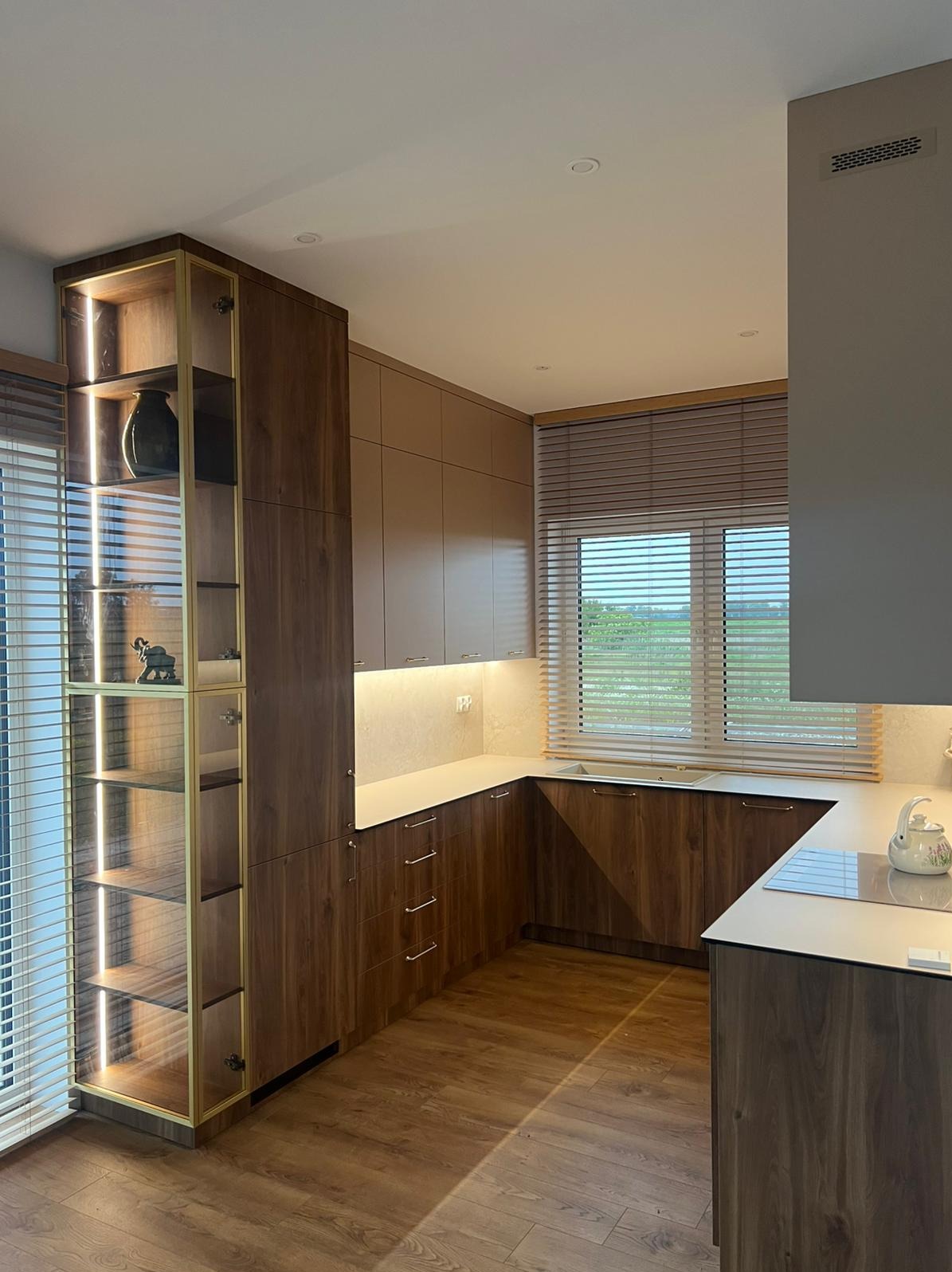 Nowoczesna kuchnia z drewnianymi frontami, oświetleniem LED w witrynie i jasnym blatem. Widok na okno z żaluzjami.