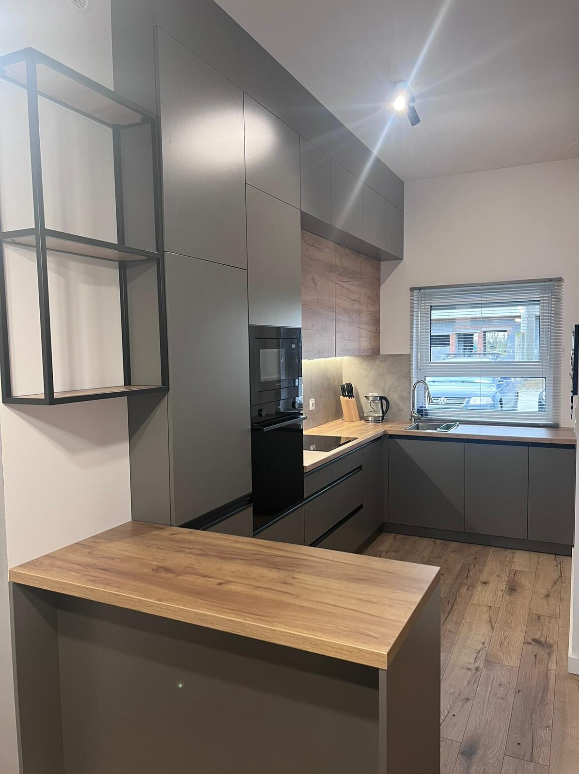 Nowoczesna kuchnia z szarymi frontami i drewnianymi akcentami, oświetleniem punktowym i wyspą kuchenną z blatem w kolorze drewna.