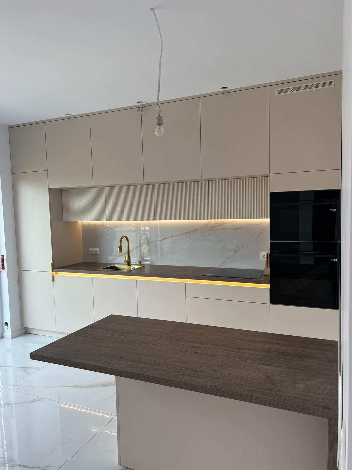 Nowoczesna kuchnia na wymiar w beżowych odcieniach z wyspą, złotą armaturą i marmurowym backsplashiem. Minimalistyczny design z oświetleniem LED.