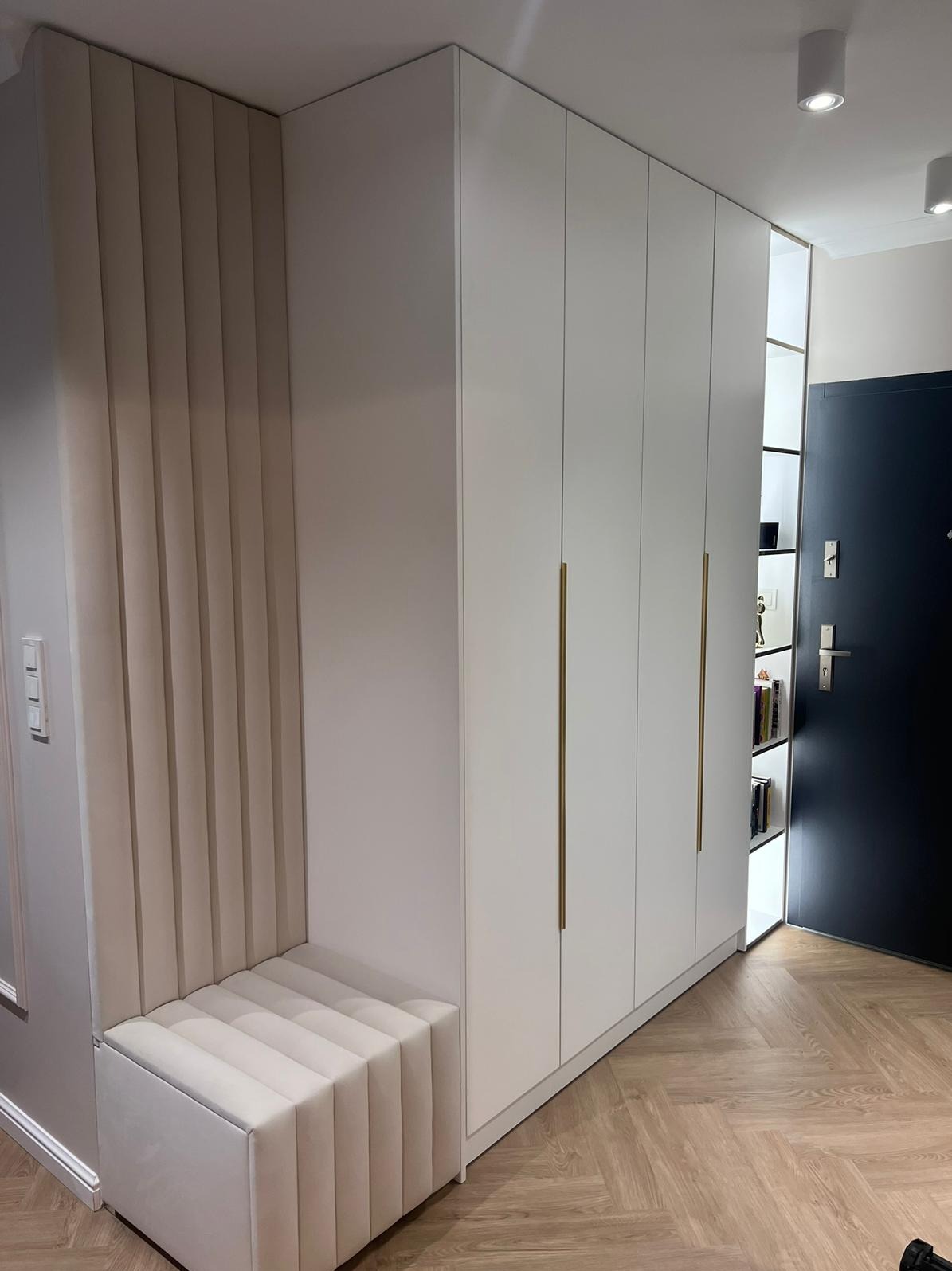 Biała szafa wnękowa z siedziskiem obitym beżową tkaniną w przedpokoju. Złote uchwyty, parkiet w jodełkę i czarne drzwi wejściowe.