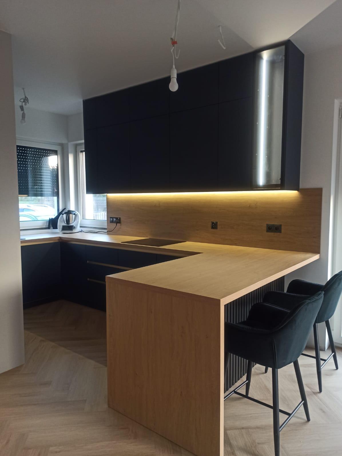 Nowoczesna kuchnia z czarnymi szafkami wiszącymi i drewnianym blatem, oświetlona listwą LED. Dwa krzesła barowe przy wyspie. Parkiet w jodełkę na podłodze.