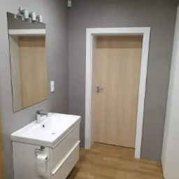 Nowoczesna łazienka z szarą ścianą, białą szafką umywalkową z dwiema szufladami, lustrem z oświetleniem LED i drewnopodobną podłogą, widoczny fragment toalety w dolnym lewym rogu.