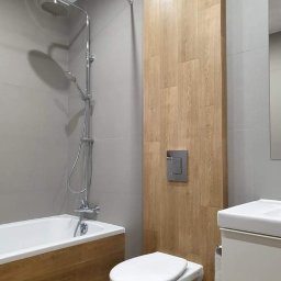 Wojciech Żaborowski - Nowoczesna łazienka z szarą glazurą i drewnopodobnymi akcentami: obudowa wanny i pionowy pas płytek imitujących drewno. Widoczna bateria prysznicowa, toaleta podwieszana i umywalka z szafką.