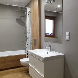 Wojciech Żaborowski - Nowoczesna łazienka z drewnianymi akcentami: obudowa wanny i panel ścienny, szare płytki, biała umywalka z szafką, lustro z oświetleniem punktowym, kabina prysznicowa z zasłoną w czarne krople.