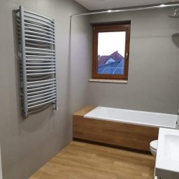 Wojciech Żaborowski - Jasna łazienka z drewnopodobną podłogą, grzejnikiem drabinkowym na ścianie, zabudowaną wanną z drewnianą obudową i oknem z widokiem na dom z panelami słonecznymi.