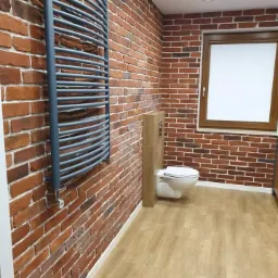 Łazienka z ceglaną ścianą, drewnianą podłogą i grzejnikiem drabinkowym w kolorze szarym, widoczne sanitariaty i okno z drewnianą ramą.