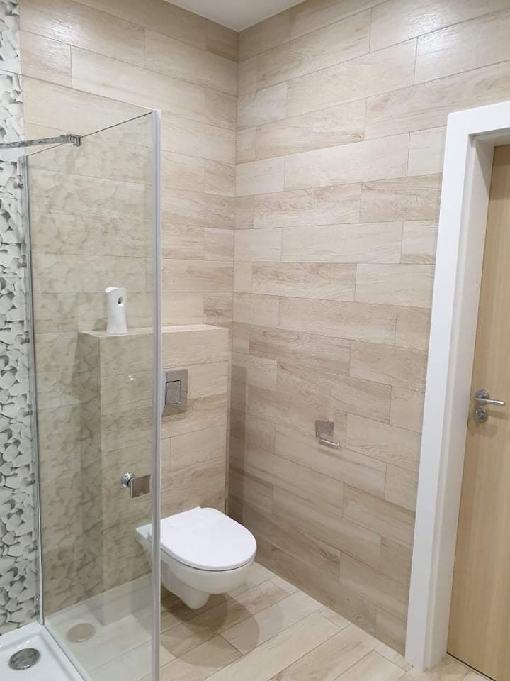 Nowoczesna łazienka z kabiną prysznicową z dekoracyjną ścianą, podwieszaną toaletą i płytkami imitującymi drewno na ścianach i podłodze, uzupełniona o białe detale.