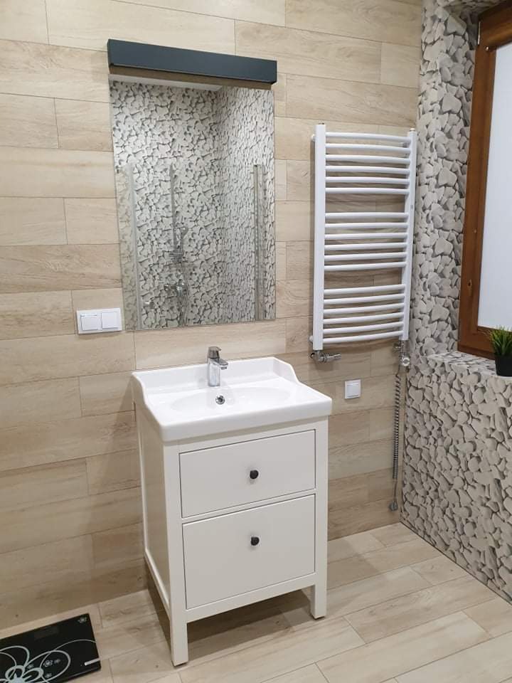 Biała szafka umywalkowa z dwoma szufladami w łazience z beżowymi płytkami imitującymi drewno i ścianą wykończoną płytkami imitującymi kamień, obok grzejnik drabinkowy