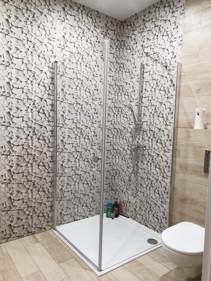 Wykończona łazienka z kabiną prysznicową, ściany wyłożone tapetą imitującą kamień, podłoga z paneli imitujących drewno, widoczny fragment toalety i dozownik zapachu.