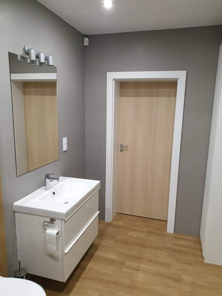 Nowoczesna łazienka z szarą ścianą, białą szafką umywalkową z dwiema szufladami, lustrem z oświetleniem LED i drewnopodobną podłogą, widoczny fragment toalety w dolnym lewym rogu.